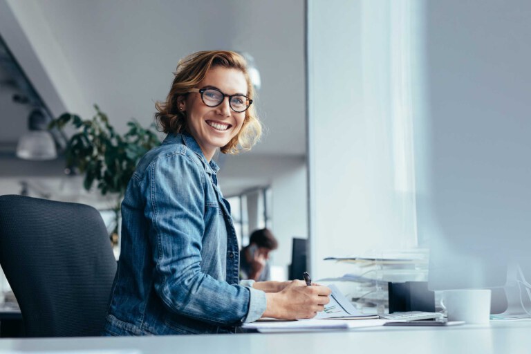 Lächelnde Frau mit Brille und Jeansjacke sitzt mit einem Stift am Schreibtisch in einem hellen Büro