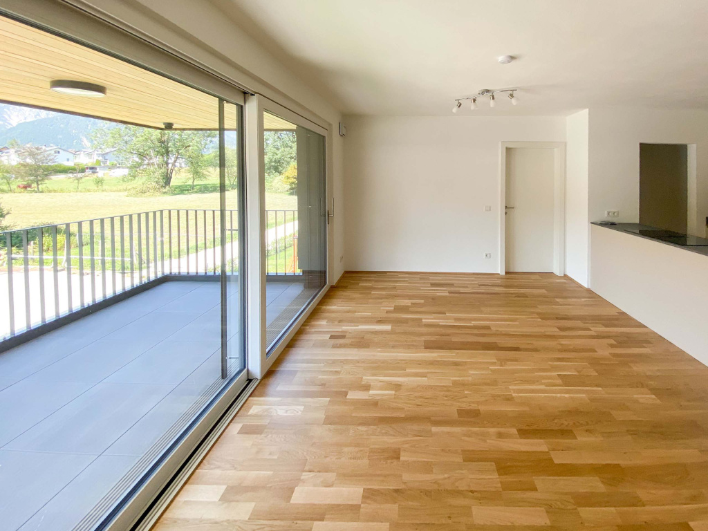 Leerer Wohnraum mit Parkettboden, offener Küche und großer Glasfront zur überdachten Terrasse