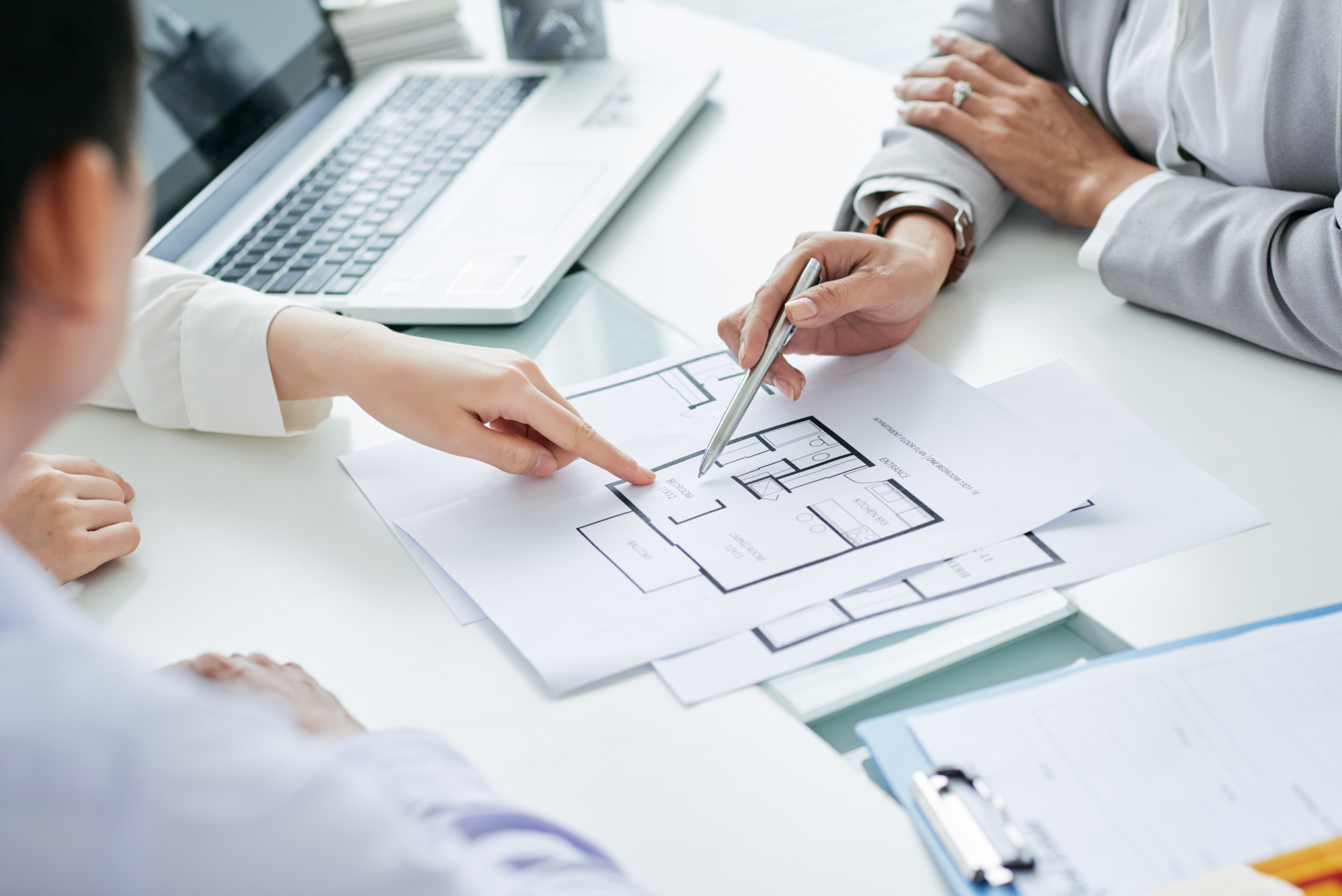 Mehrere Hände arbeiten an Laptops und zeigen auf Baupläne neben einem weißen Architekturmodell
