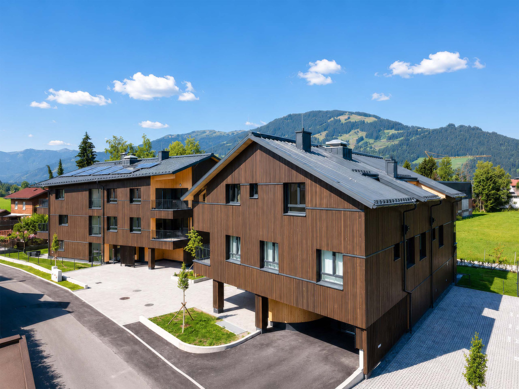 Zwei moderne Wohnhäuser mit Holzfassaden vor bewaldeten Bergen unter blauem Himmel