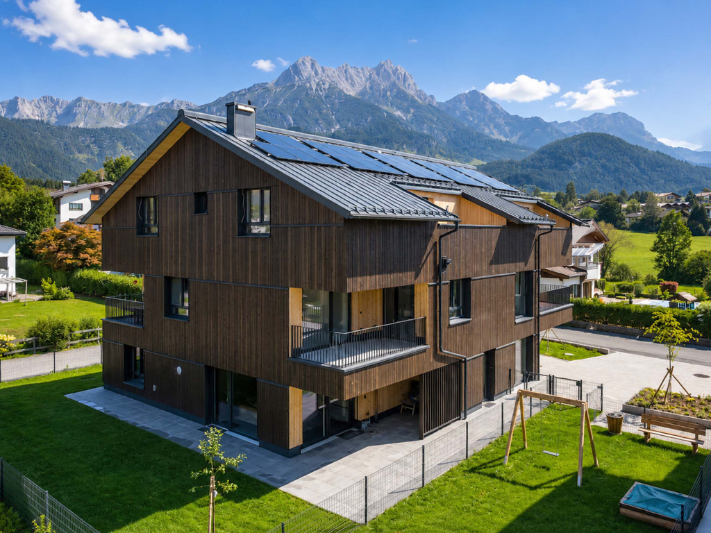 Mehrstöckiges Wohnhaus mit dunkler Holzfassade, Solarpaneelen und Balkonen vor einer Bergkulisse