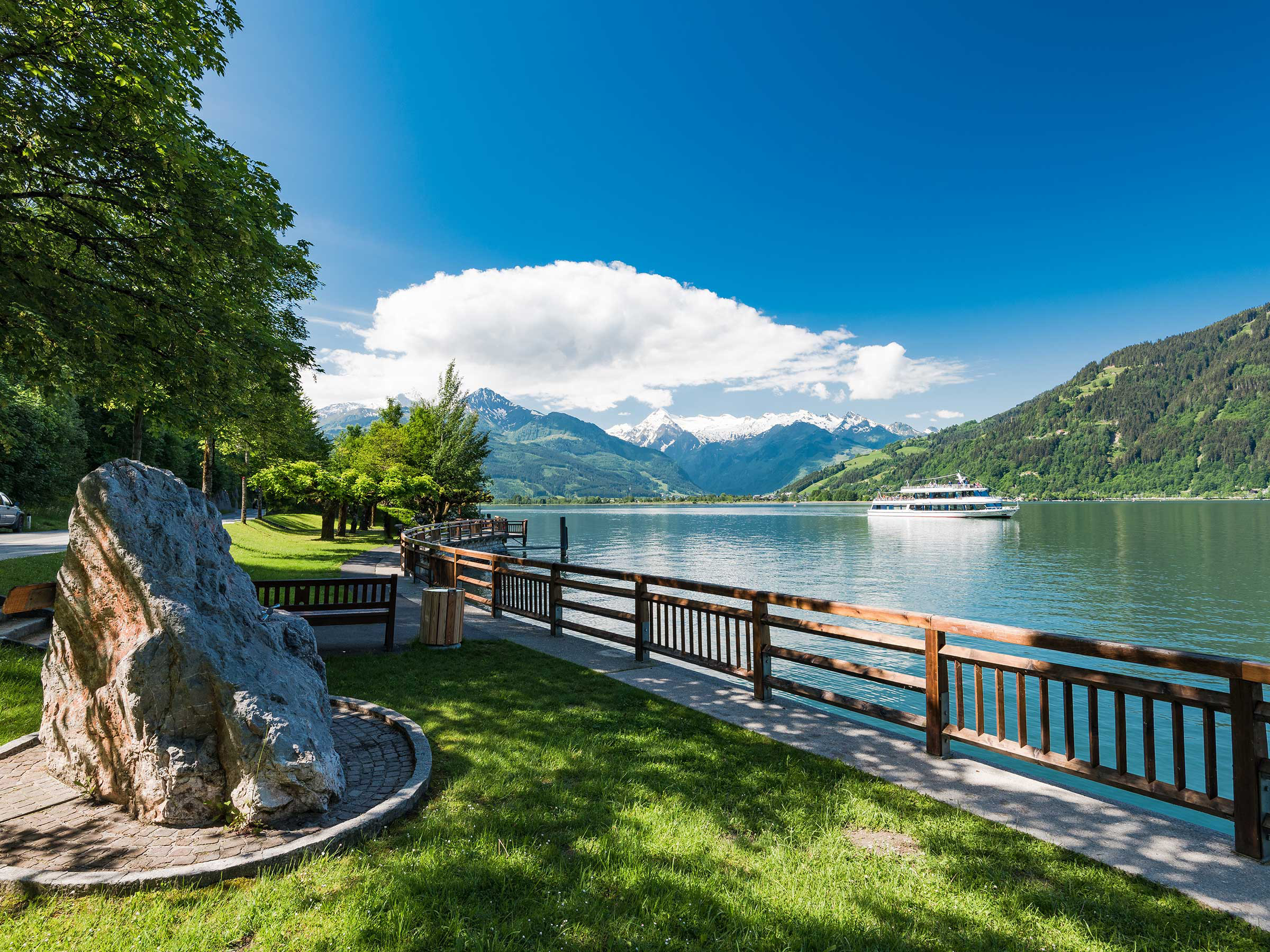 Uferweg am See Thumersbach mit Ausflugsschiff, grünen Bergen und schneebedeckten Gipfeln